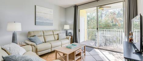 A living room with a sofa, coffee table, and a TV on a stand. There is a balcony with a table and chairs.