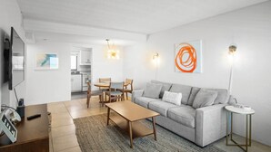 A modern living room with a sofa, coffee table, and dining area. There is a TV, a painting, and a lamp on the wall. - Oceanfront Daytona Beach Condo w/ Beach Views! (Daytona Beach)