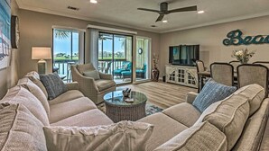 A living room with a large sectional sofa, a glass coffee table, and a television. There is a ceiling fan, a clock, and a wall-mounted sign. - Beachy Lakefront Escape - Walk to the Shore! (Miramar Beach)