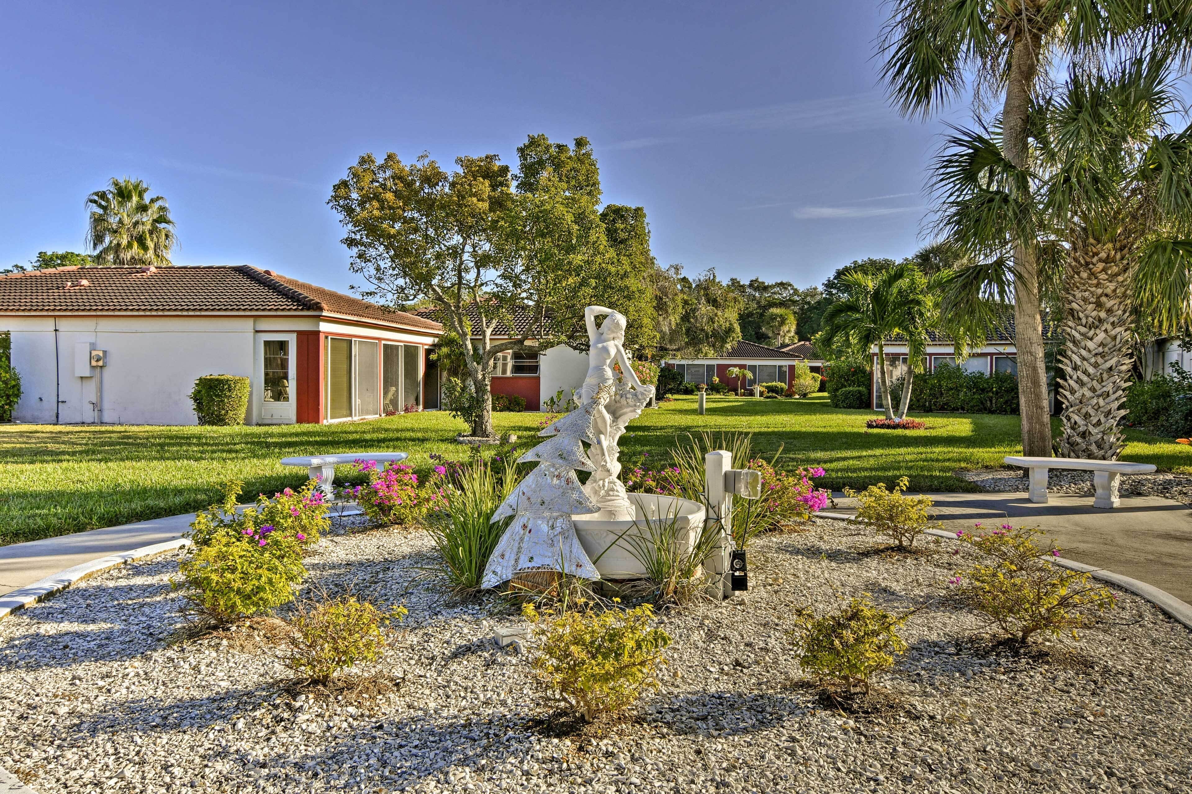 A sculpture garden with a prominent white sculpture, surrounded by gravel, small shrubs, and flowering plants.