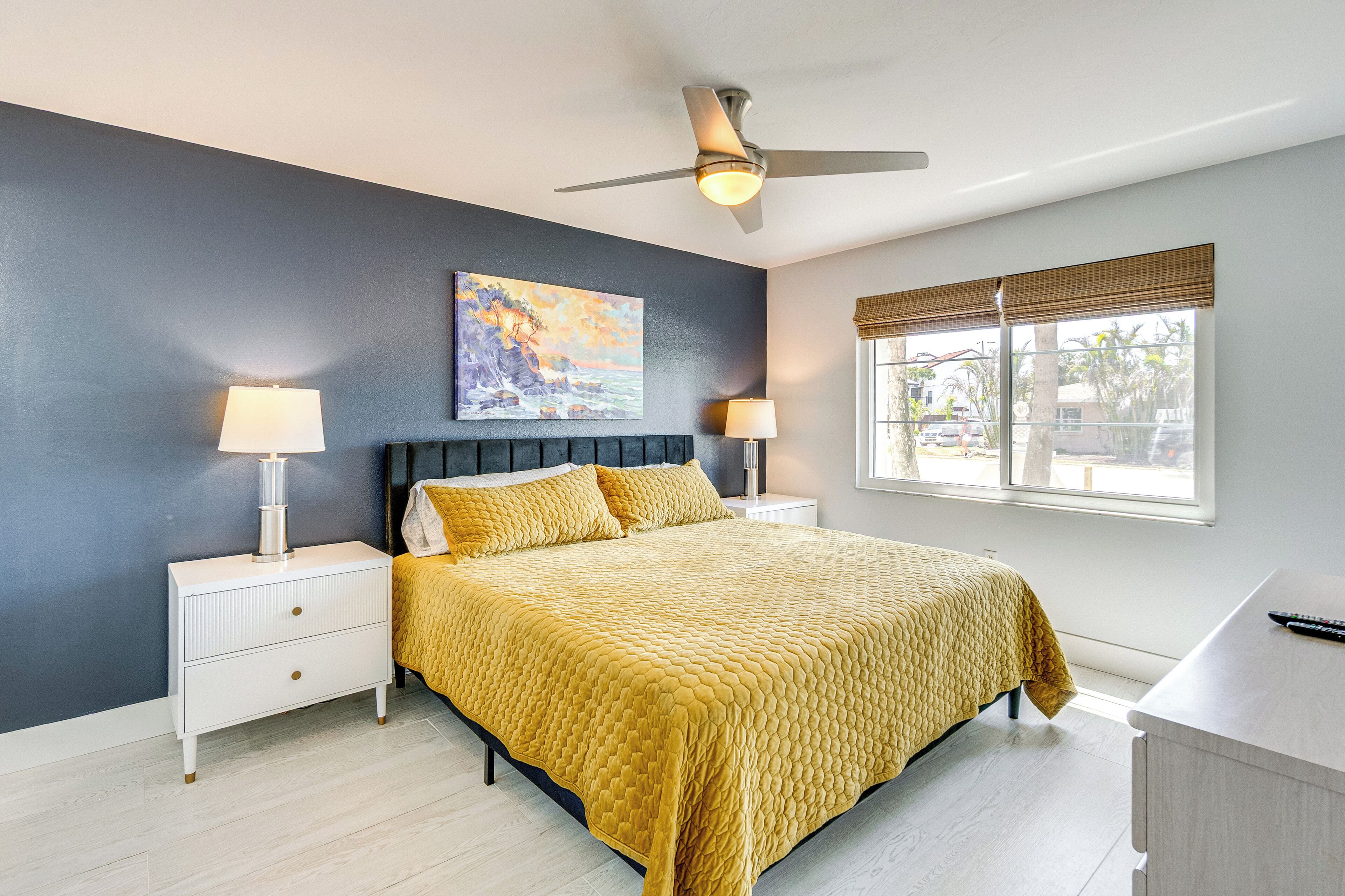 A bedroom with a bed, two bedside tables, a ceiling fan, a window with blinds, and a painting on the wall.