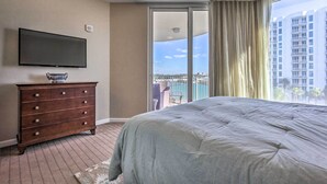Ett hotellrum med en stor säng, en platt-TV som är monterad på väggen, en byrå i trä och en skjutdörr i glas som leder ut till en balkong med utsikt över höga byggnader och en pool.