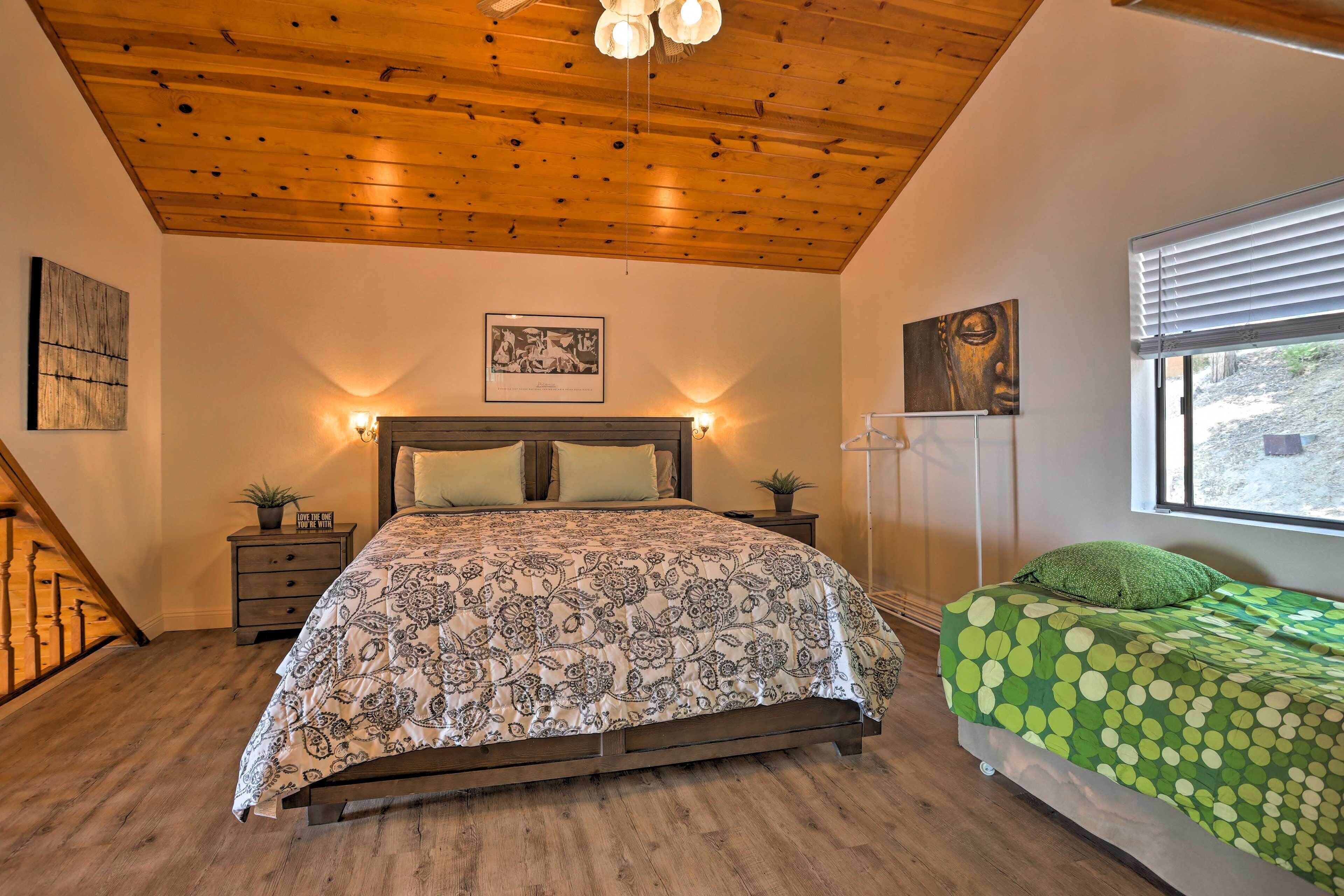 A bedroom with a wooden ceiling, a bed with patterned bedding, a green bed on the right, and a window with blinds.