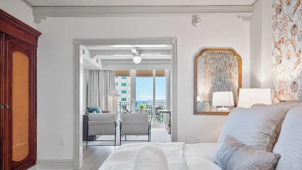 A hotel room with a bed, a nightstand, a window with curtains, a balcony with a view of buildings, and a door leading to another room. - Sunny Hammock Beach Condo: Balcony w/ Ocean Views! (Palm Coast)
