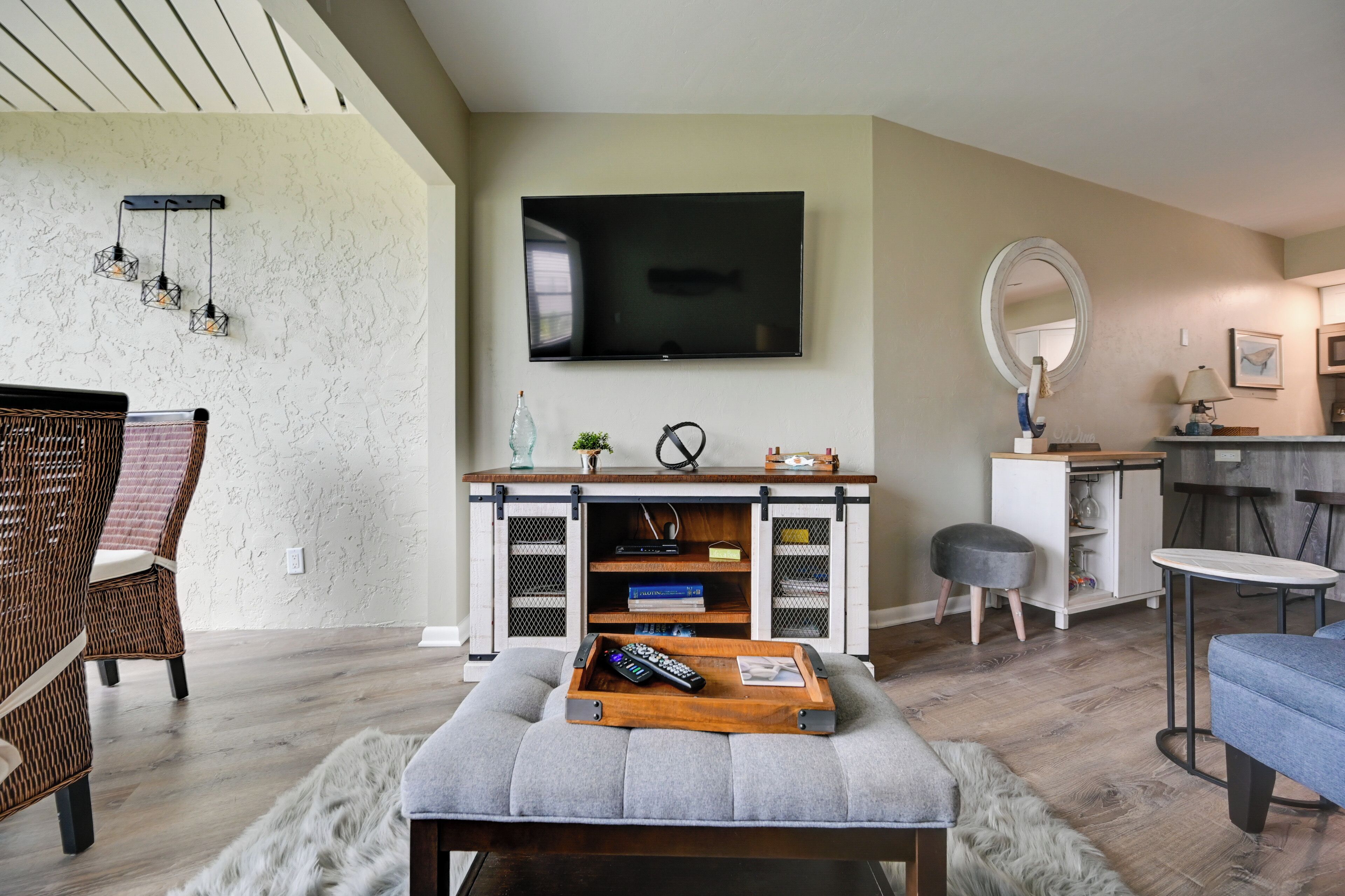 Una sala de estar con televisor de pantalla plana, una consola blanca con libros, un otoman gris, sillas de mimbre y un comedor con un espejo redondo y taburetes de bar.
