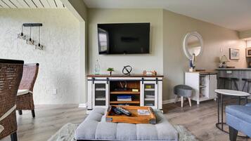 Una sala de estar con televisor de pantalla plana, una consola blanca con libros, un otoman gris, sillas de mimbre y un comedor con un espejo redondo y taburetes de bar.