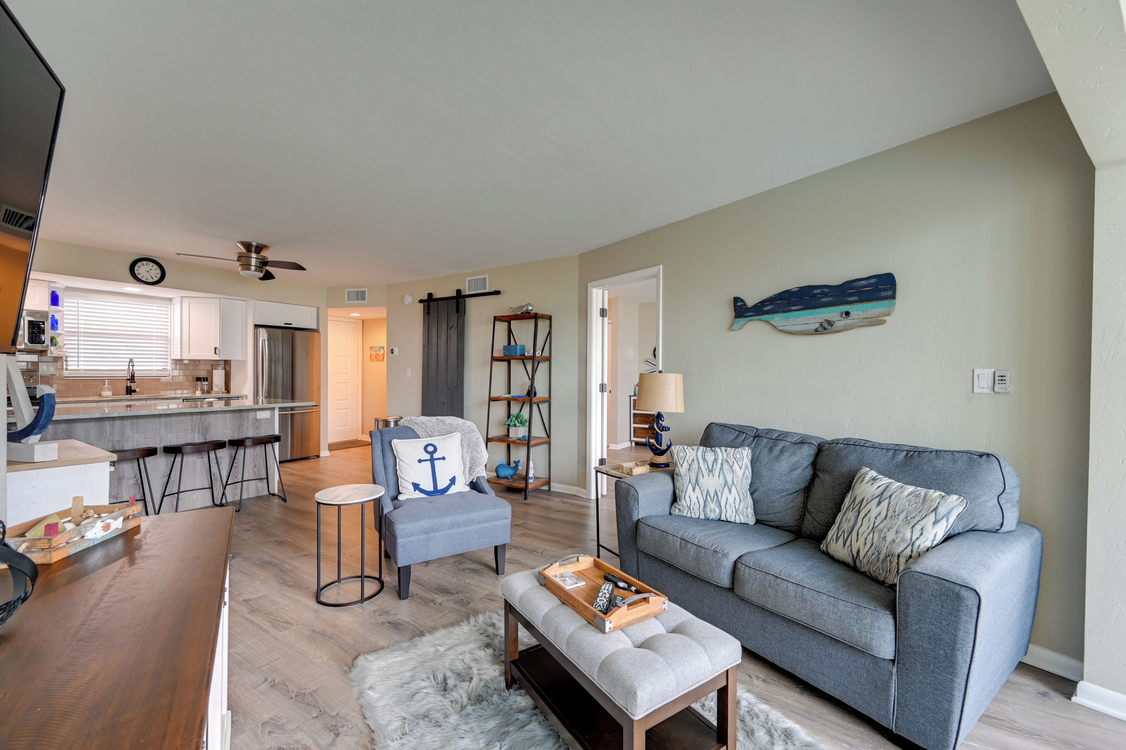 Una sala de estar moderna con un sofá gris, una mesa de centro y una mesita auxiliar. Hay una zona de cocina con un refrigerador y una mesa de comedor.