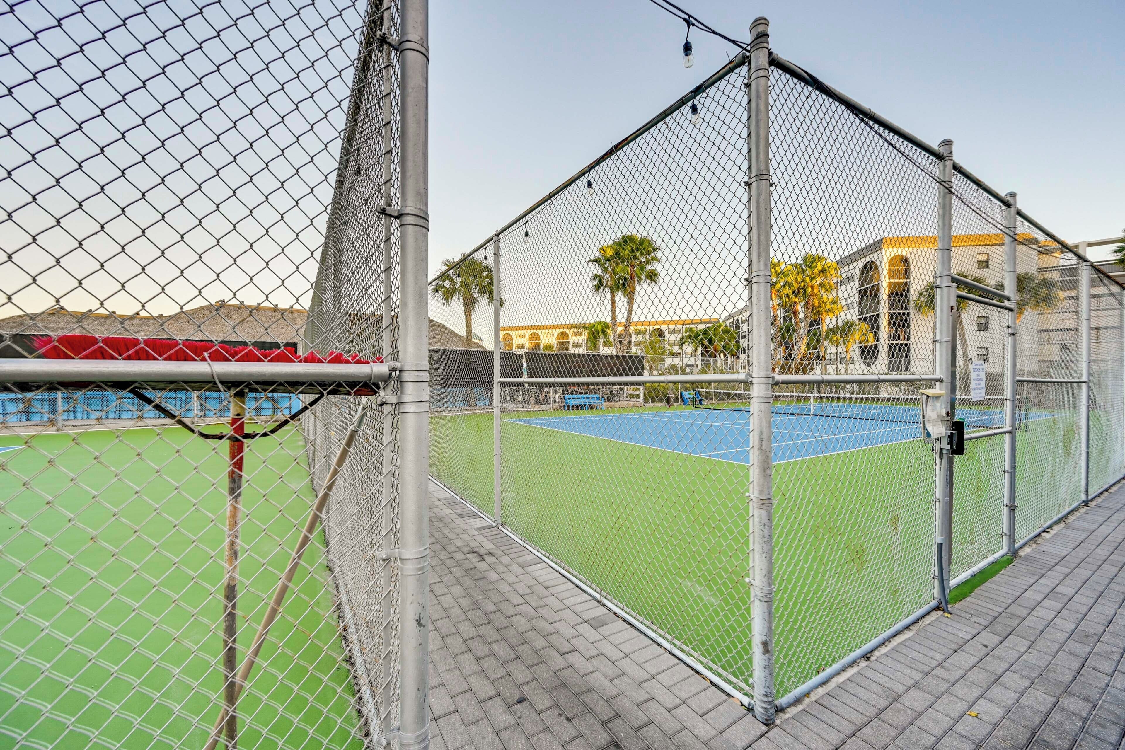 Una cancha de tenis cercada con vista despejada del entorno, incluyendo un edificio de fachada amarilla y palmeras.