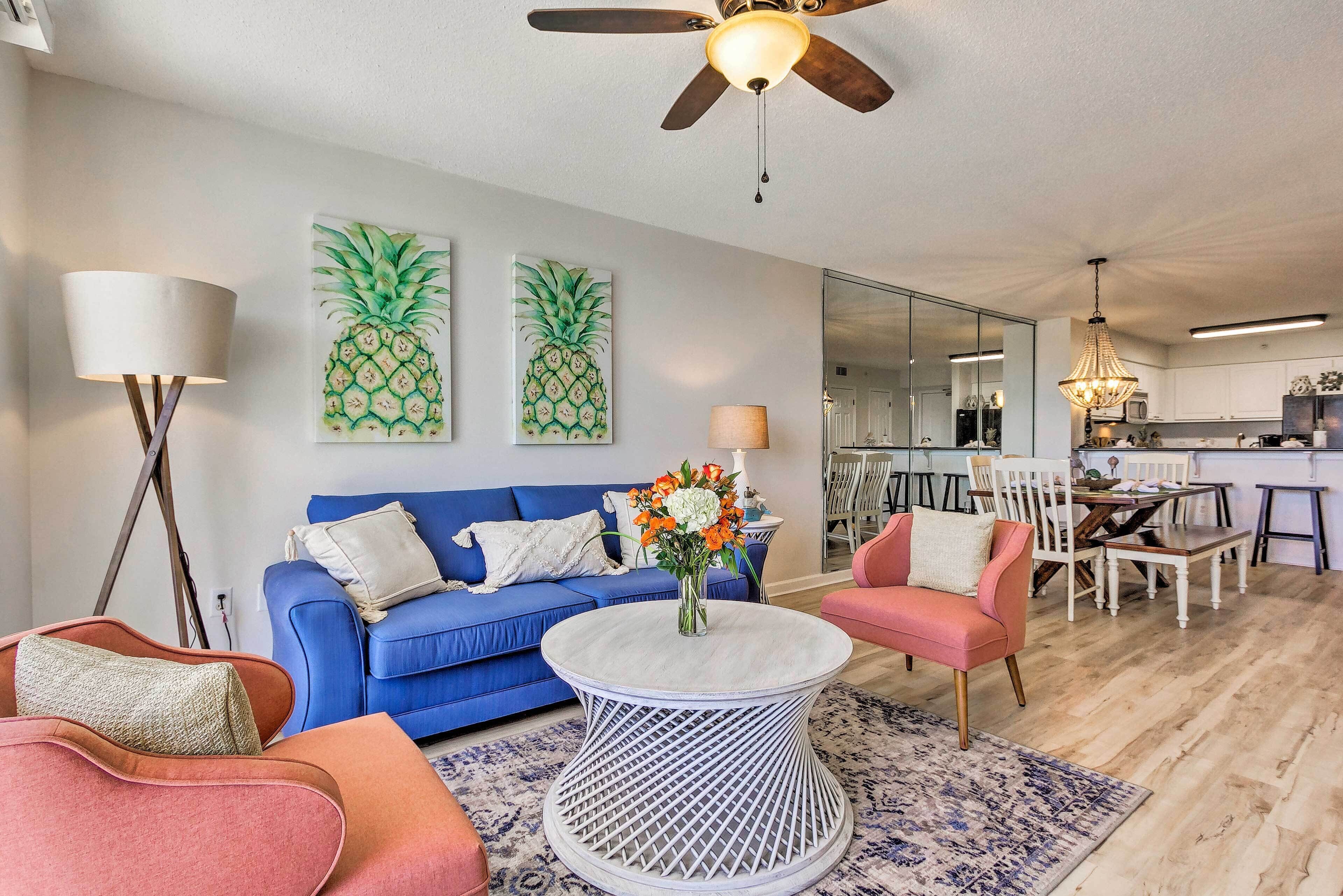 A modern living room with a blue sofa, a pink armchair, and a round coffee table. There are two pineapple paintings on the wall, a ceiling fan, and a chandelier.