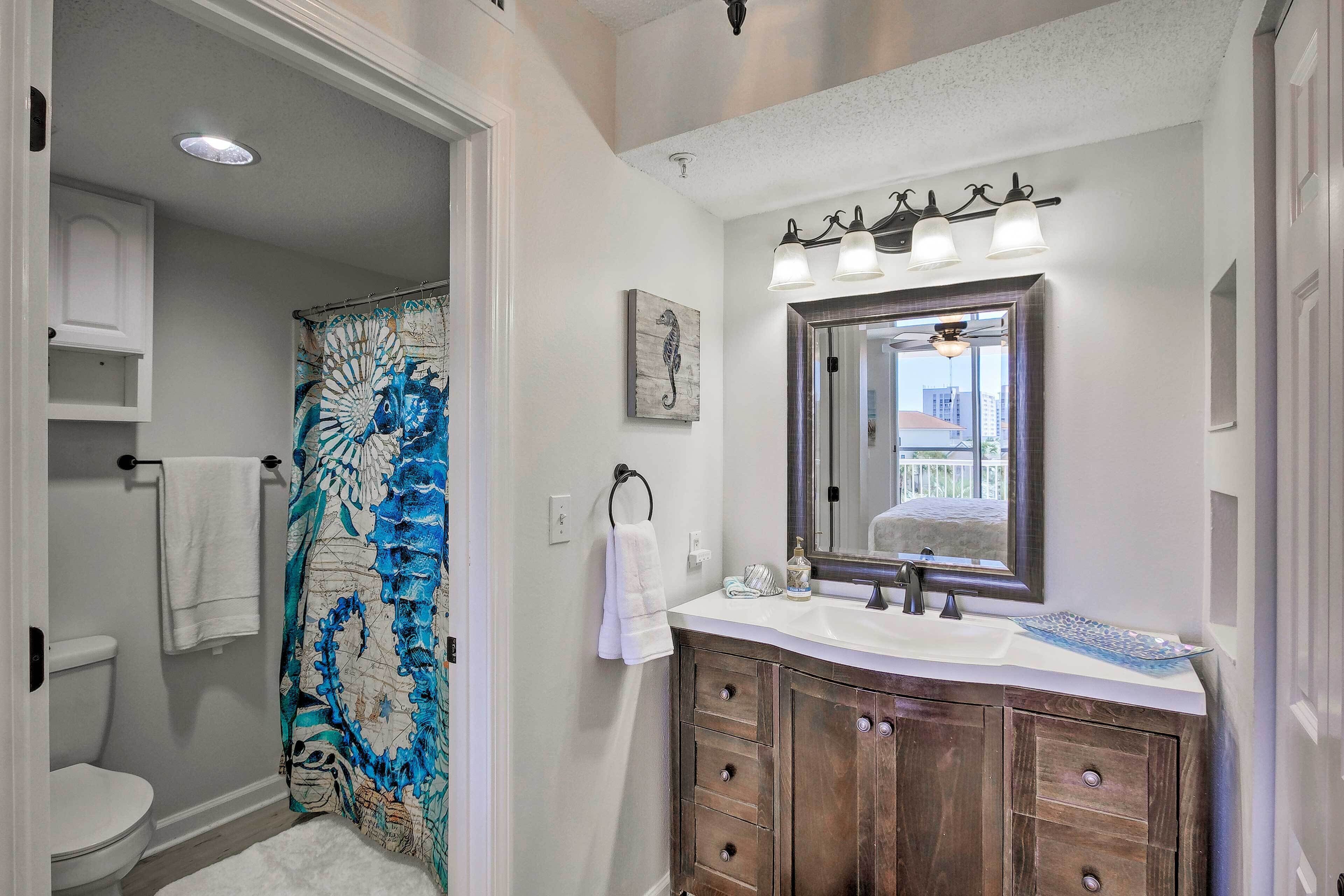 Une salle de bain avec des toilettes blanches, une douche avec un rideau de douche bleu et blanc, un meuble-lavabo en bois avec un miroir et un lavabo blanc.