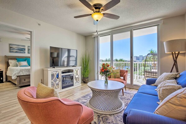 A modern living room with a ceiling fan, a flat-screen TV, a sofa, a coffee table, and a balcony with a view of the city. - Snowbird's Retreat: Walkable Destin Condo w/ View! (Destin)