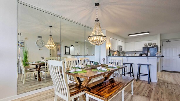 A dining area with a wooden table and chairs, a chandelier, and a kitchen in the background.