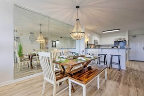 A dining area with a wooden table, white chairs, and a chandelier. There is a kitchen in the background with white cabinets and a refrigerator.