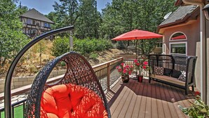 A wooden deck with a wicker chair and a red umbrella, surrounded by greenery and a house in the background.