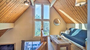A loft-style room with a sloped ceiling, wooden beams, a large window, a sofa, a wooden table, and a hanging light fixture.
