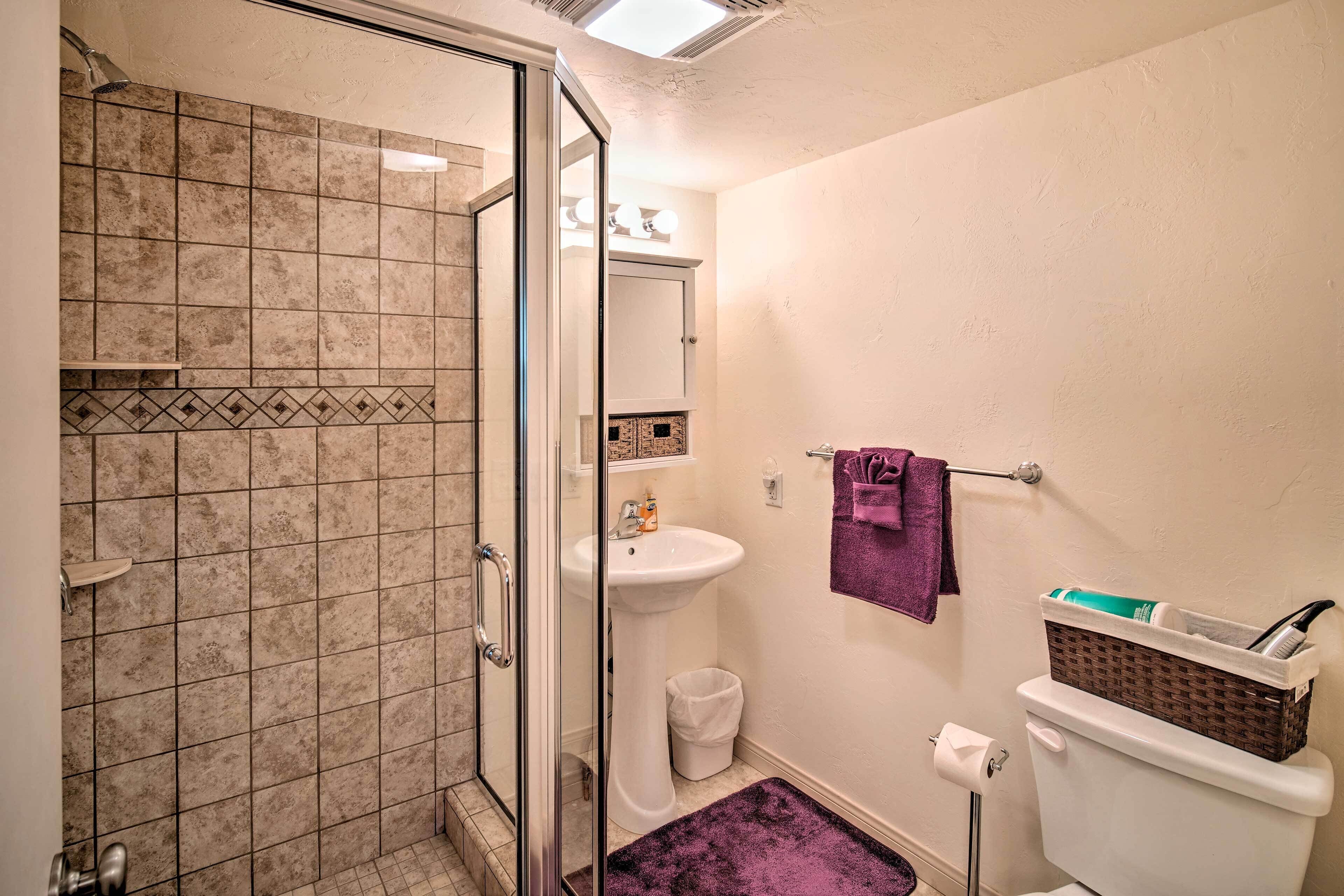 Un baño con mampara de vidrio, un lavamanos blanco, un inodoro y una alfombra de baño morada.