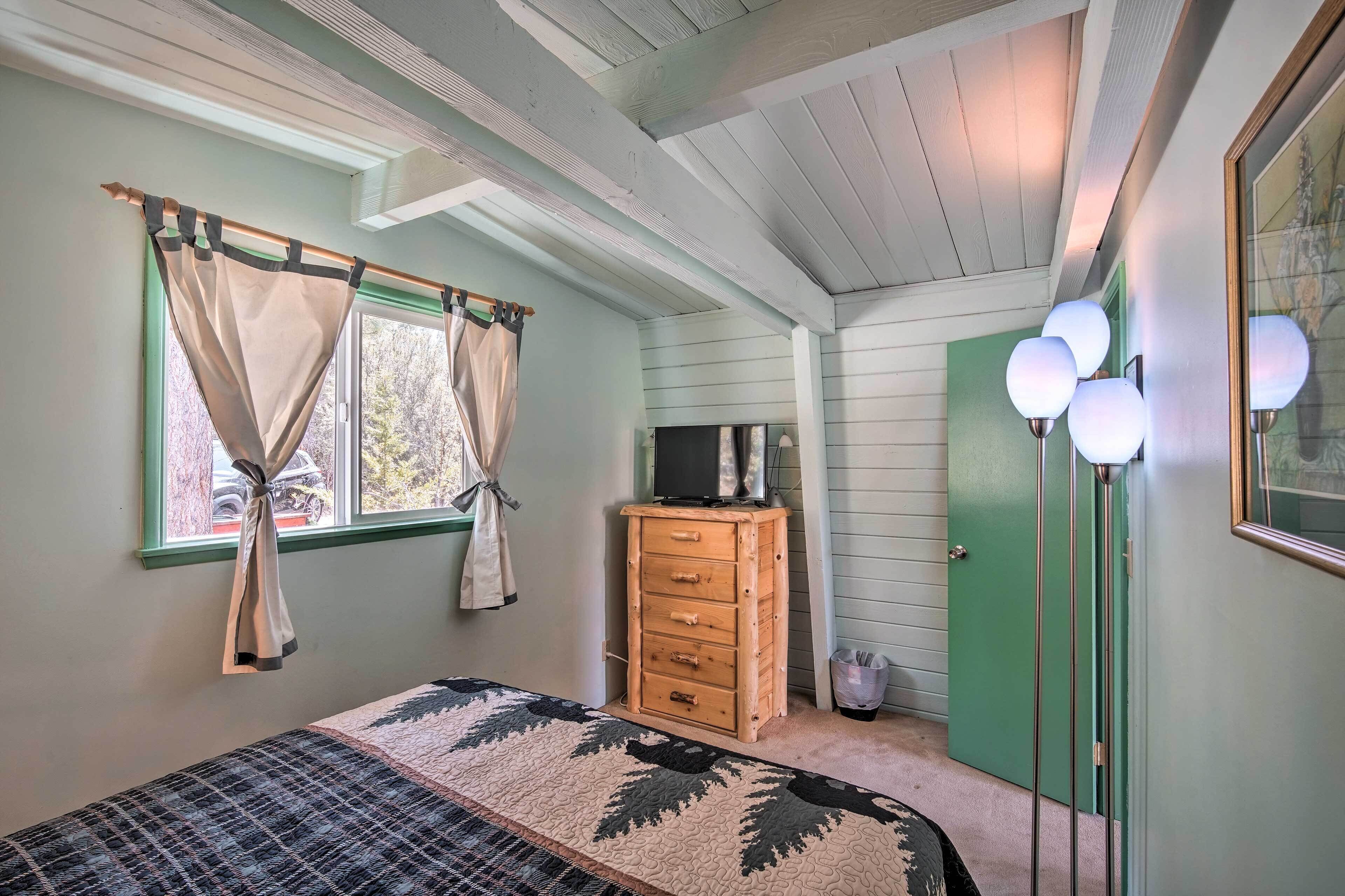 Un dormitorio con cama, cómoda, televisor encima y una puerta verde, además de una ventana con cortinas.