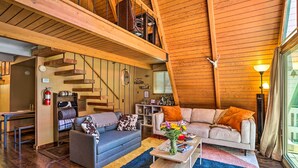 A spacious loft-style living area with a high ceiling, wooden beams, a staircase, a sofa, a coffee table, and a view through large windows.