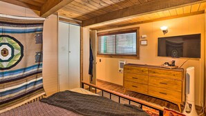 A bedroom with a wooden ceiling, a bed with patterned bedding, a dresser, a fan, and a TV on the wall.