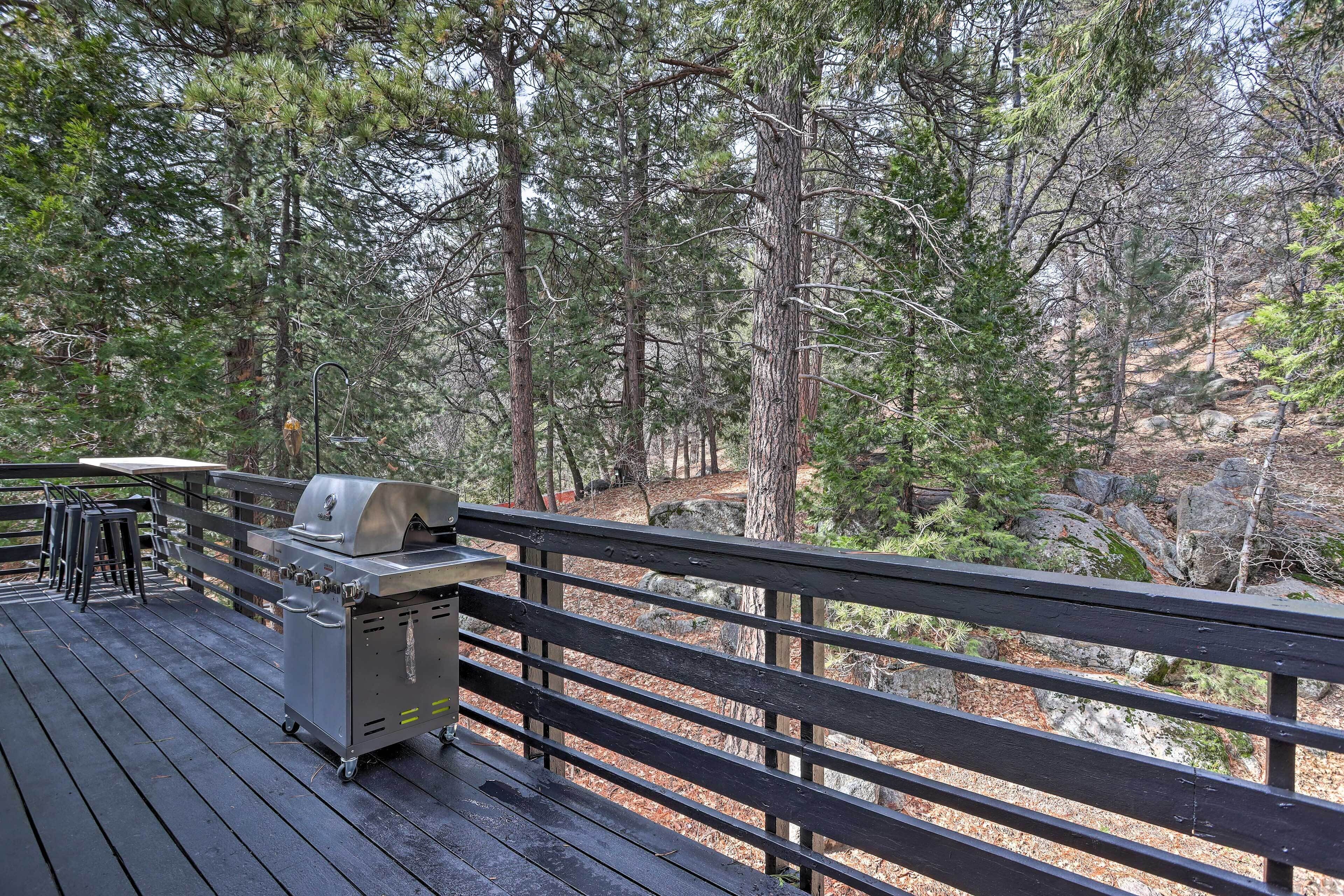 A deck with a grill and chairs, surrounded by a forested area.