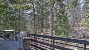 A deck with a grill and chairs, surrounded by a forested area.