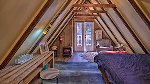 A cozy attic bedroom with exposed wooden beams, a fireplace, a bed, and a small table. - Cozy, Pet-friendly Mtn Cabin: 1 Mi to Lake Gregory (Crestline)
