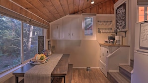 A cozy cabin interior with wooden ceilings, a dining table with a placemat, a kitchen area with a coffee station, and a view of the forest through large windows. - Cozy, Pet-friendly Mtn Cabin: 1 Mi to Lake Gregory (Crestline)