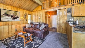 A cabin interior with a brown leather sofa, a wooden coffee table, a patterned rug, a kitchen area with a stove, and a framed picture of deer on the wall. - 5 Mi to Big Bear Mtn Resort: Cozy Cabin w/ Deck (Big Bear City)