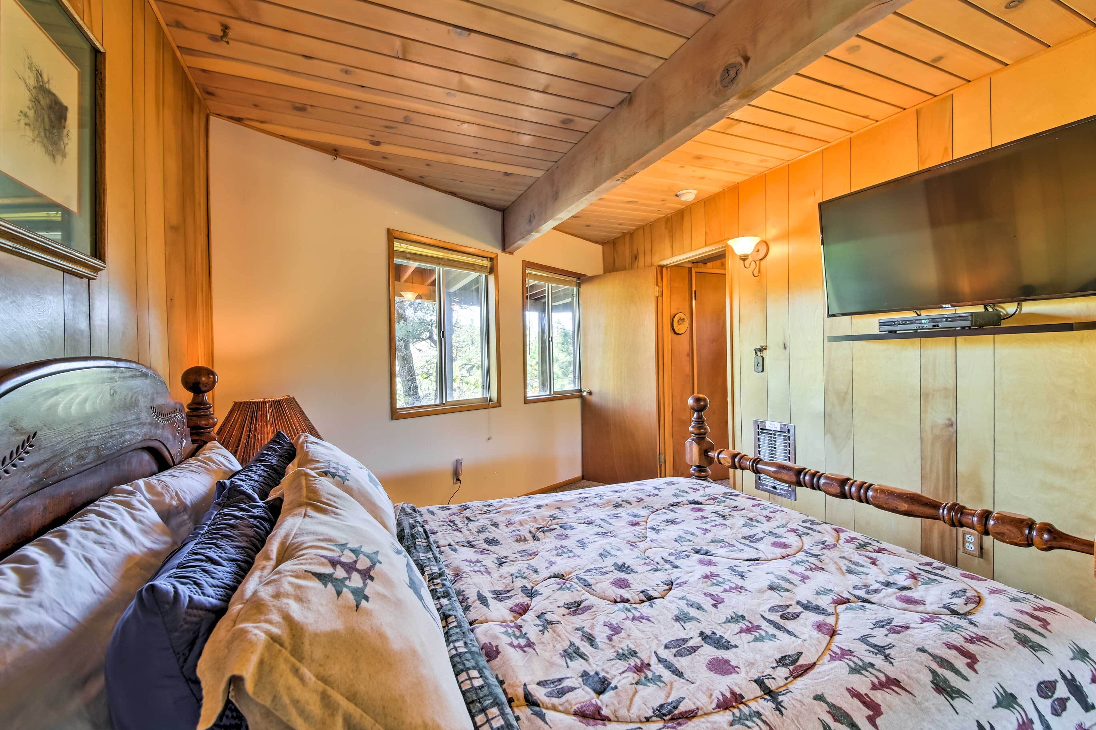 A bedroom with a wooden ceiling, a bed with a floral bedspread, a mounted TV, and a window with a view of trees.