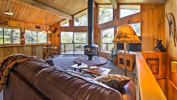 A cozy cabin interior with a fireplace, leather sofa, wooden walls, and large windows. - 5 Mi to Big Bear Mtn Resort: Cozy Cabin w/ Deck (Big Bear City)