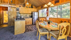 A cabin interior with wooden walls, a kitchen with a refrigerator, a dining area with a table and chairs, and a large window with a view of trees. - 5 Mi to Big Bear Mtn Resort: Cozy Cabin w/ Deck (Big Bear City)