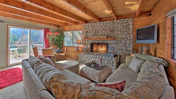 A cozy living room with a stone fireplace, wooden beams, and a large sofa. - Mountainside Cabin w/ Deck - 16 Miles to Skiing! (Lake Arrowhead)