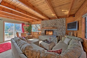 A cozy living room with a stone fireplace, wooden beams, and a large sofa. - Mountainside Cabin w/ Deck - 16 Miles to Skiing! (Lake Arrowhead)
