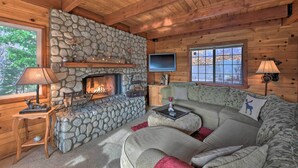 A cozy cabin interior with a stone fireplace, wooden walls, a sofa, and a television. - Mountainside Cabin w/ Deck - 16 Miles to Skiing! (Lake Arrowhead)
