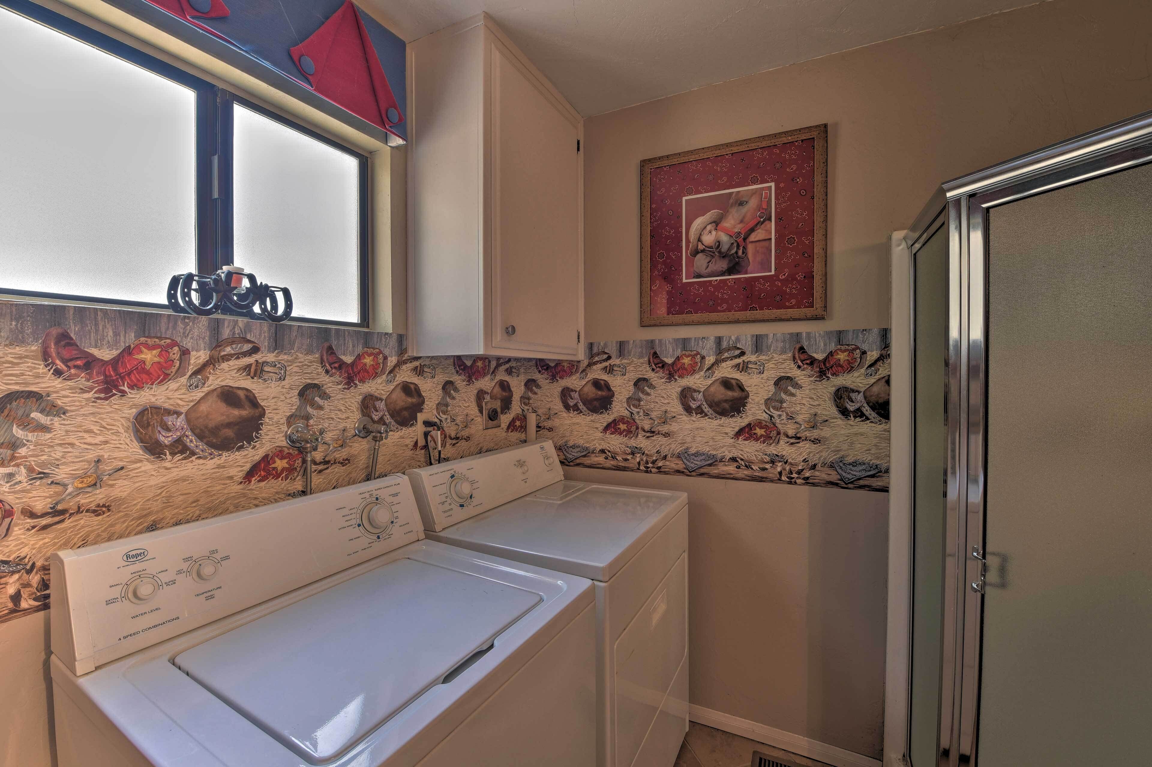 Un lavadero con dos lavadoras de carga frontal, una ventana con persianas, una pared decorativa con un cuadro en marco rojo y un armario metálico.