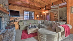 A cozy log cabin interior with a stone fireplace, a large sectional sofa, a flat-screen TV, and a staircase leading to an upper level. - Mountainside Cabin w/ Deck - 16 Miles to Skiing! (Lake Arrowhead)