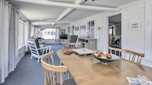 A dining area with a wooden table, chairs, and a bowl of fruit. There are white walls and a window with curtains. - Plum Island Retreat ~ 1-block Walk to Beach! (Newbury)