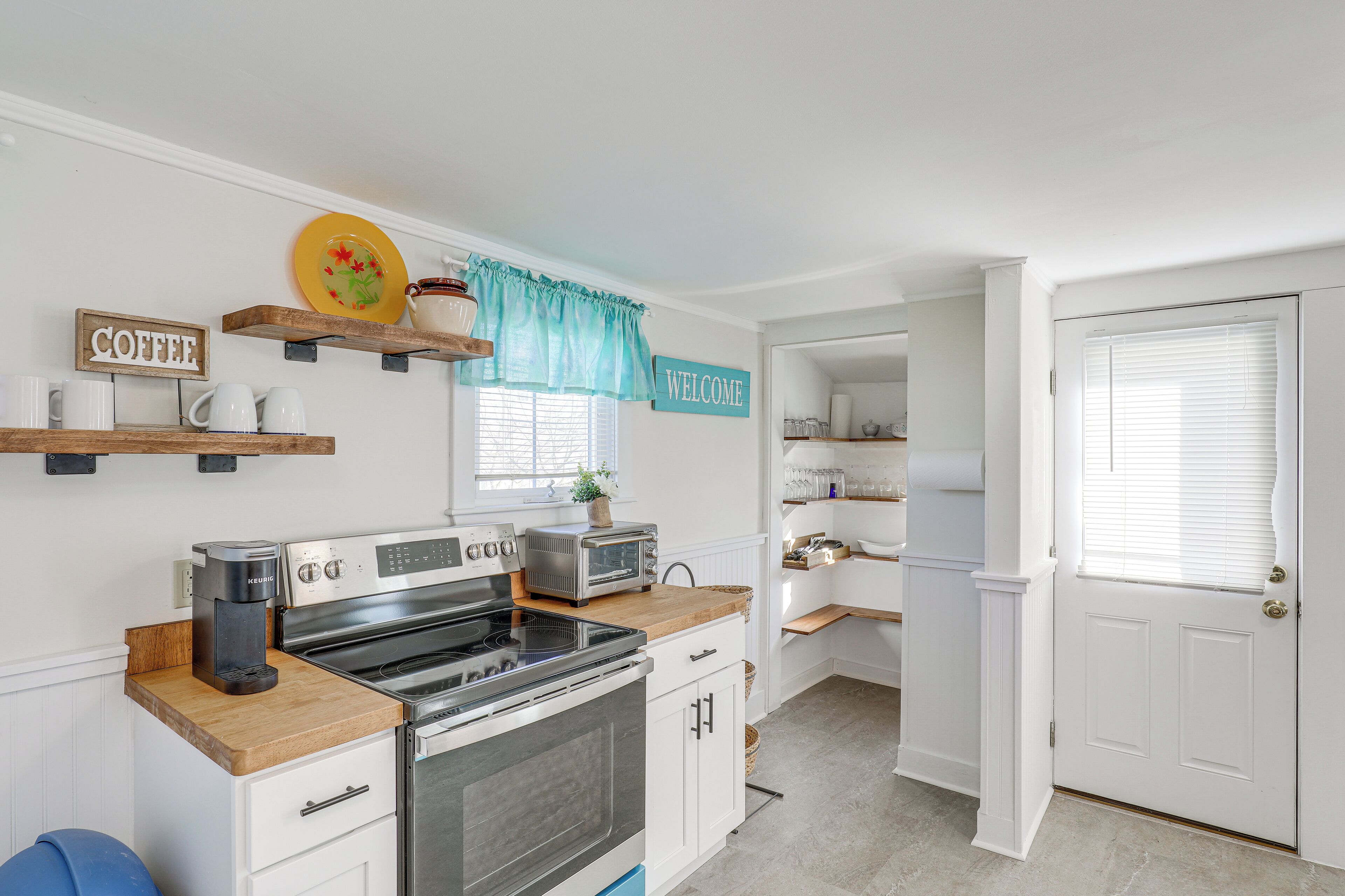 A kitchen with white cabinetry, a coffee maker, a toaster, and a stove. There are wooden shelves above the counter and a welcome sign on the wall.