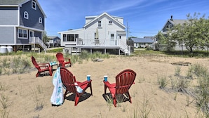 A beachside property with a large house, a covered patio with red chairs and a table, and a sandy beach area. - Plum Island Retreat ~ 1-block Walk to Beach! (Newbury)