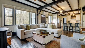 A spacious living room with a large sectional sofa, a coffee table, and a fireplace. There is a kitchen area with bar stools and a dining table. - Historic Home w/ Modern Updates on < 4 Acres (Royalston)