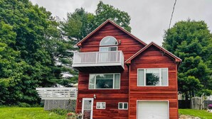 A two-story red wooden house with a balcony, a garage, and a fenced backyard. - Lakeside Berkshires Home Near Hiking & Skiing! (Lanesborough)