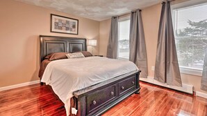 A bedroom with a large bed, a headboard with a framed picture, a nightstand, a window with curtains, and a radiator under the window.