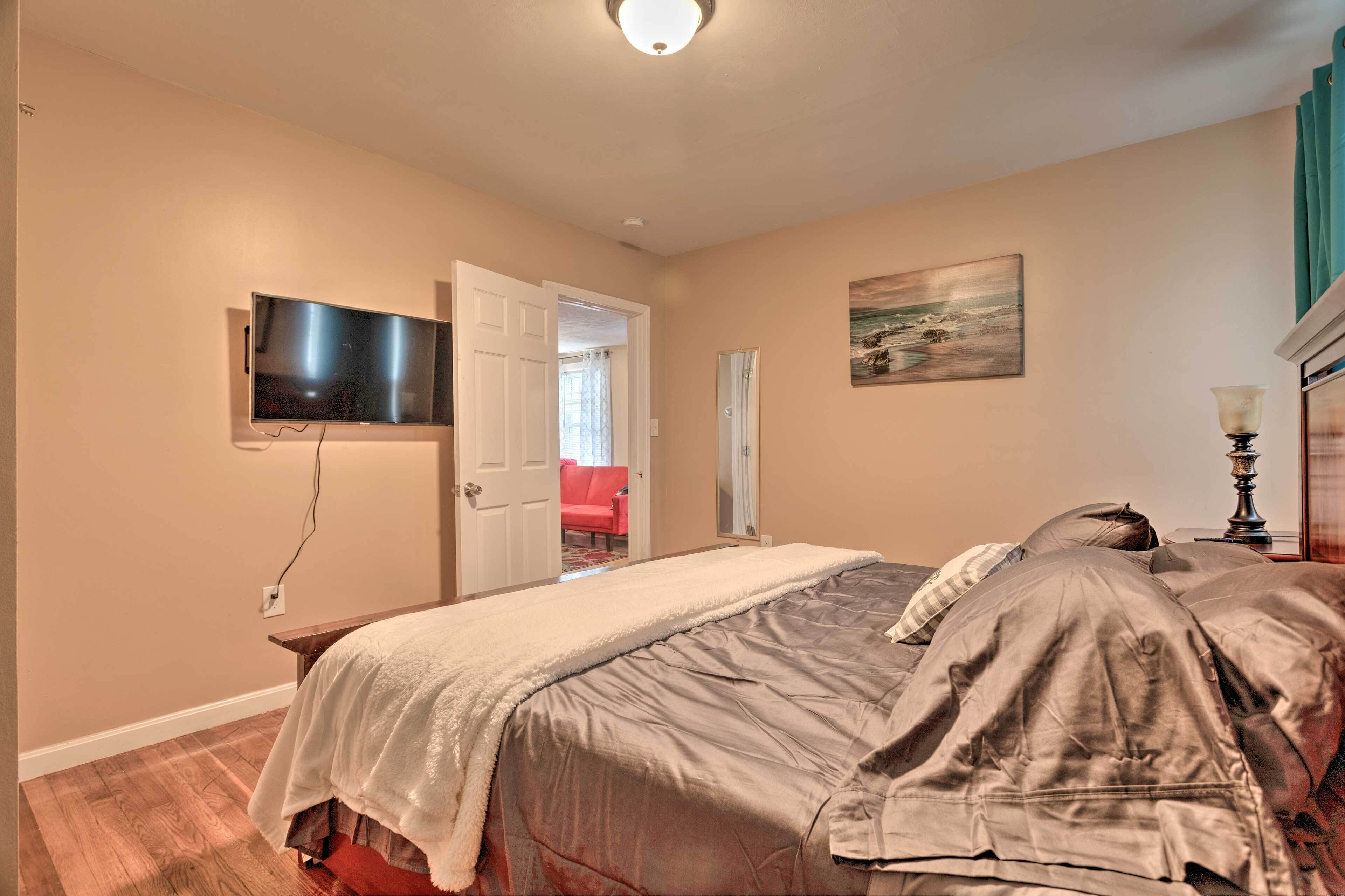 Un dormitorio con cama, televisor montado en la pared, puerta que da a otra habitación y un cuadro colgado en la pared.