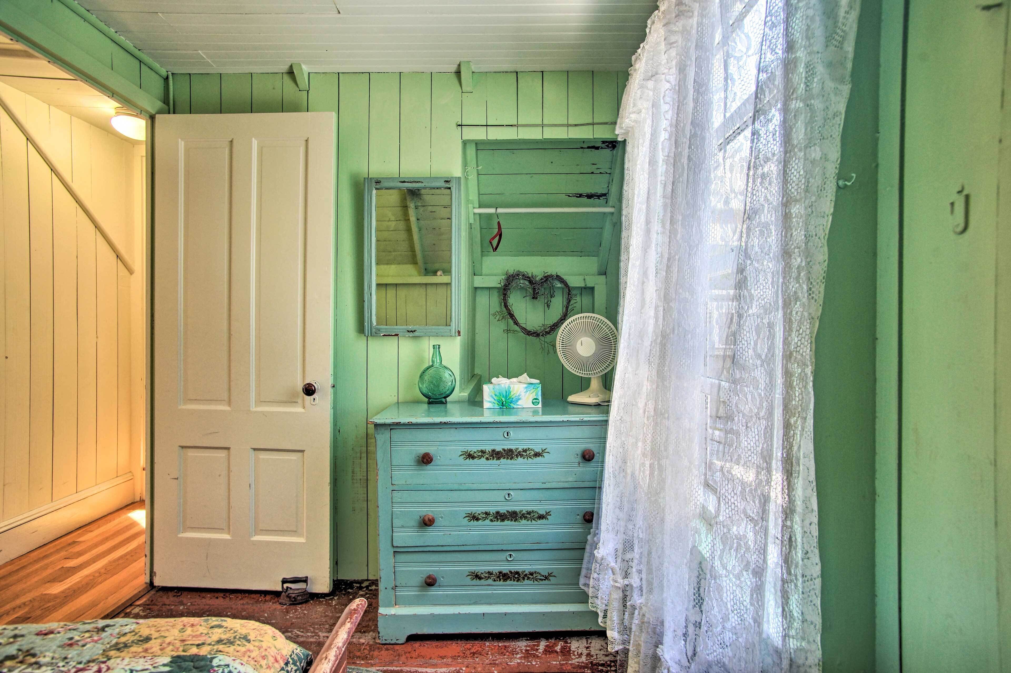 Una habitación con una pared verde, una puerta blanca, un aparador con cajones, un espejo, un ventilador y una cortina de encaje.