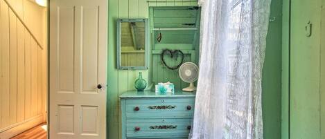 Una habitación con una pared verde, una puerta blanca, un aparador con cajones, un espejo, un ventilador y una cortina de encaje.
