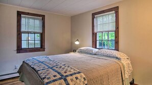 A bedroom with a large bed, two windows with blinds, a nightstand with a lamp, and a wooden floor.