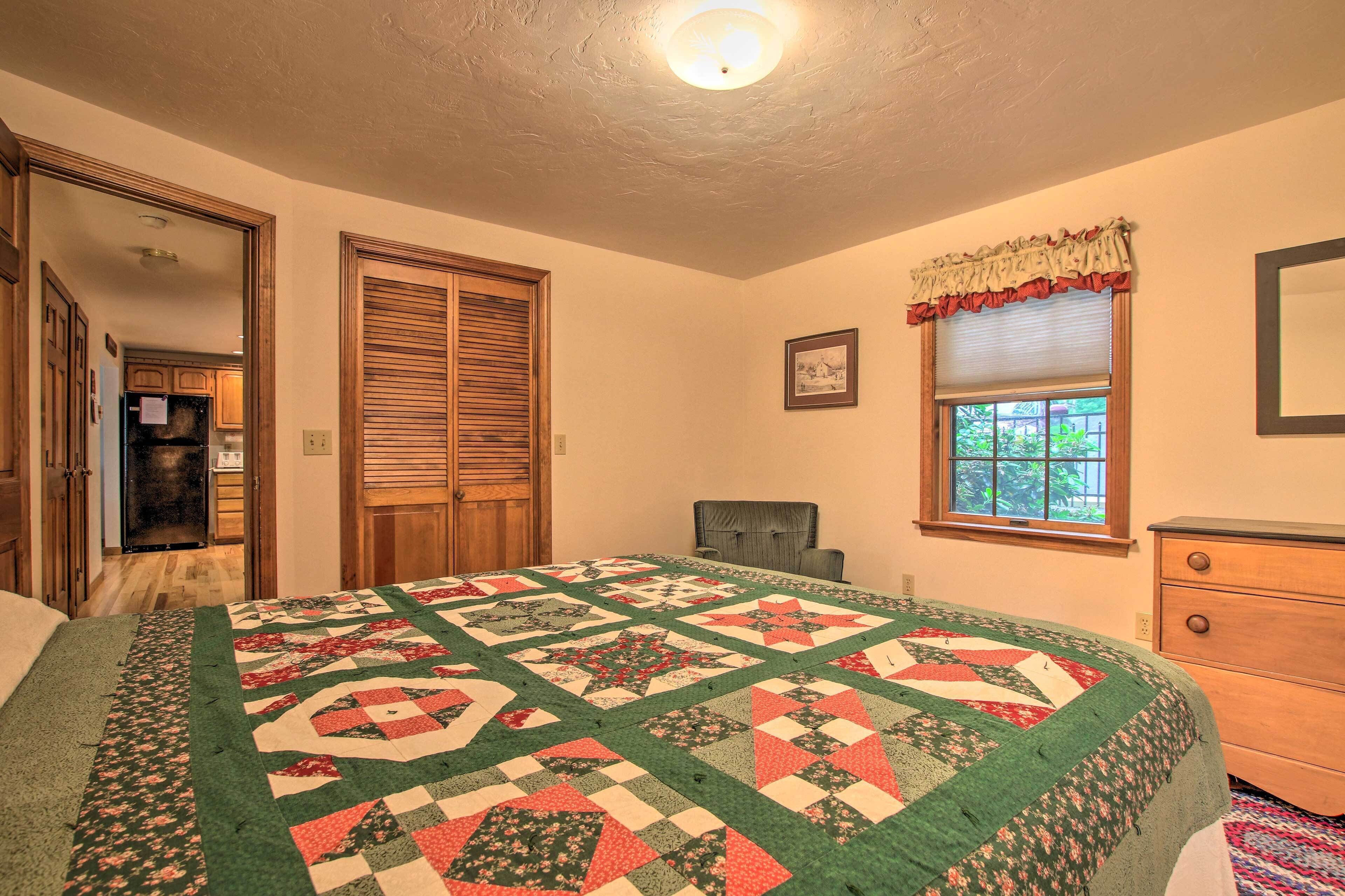 Quarto com uma cama grande, cômoda de madeira, janela com vista e porta que dá para uma cozinha.