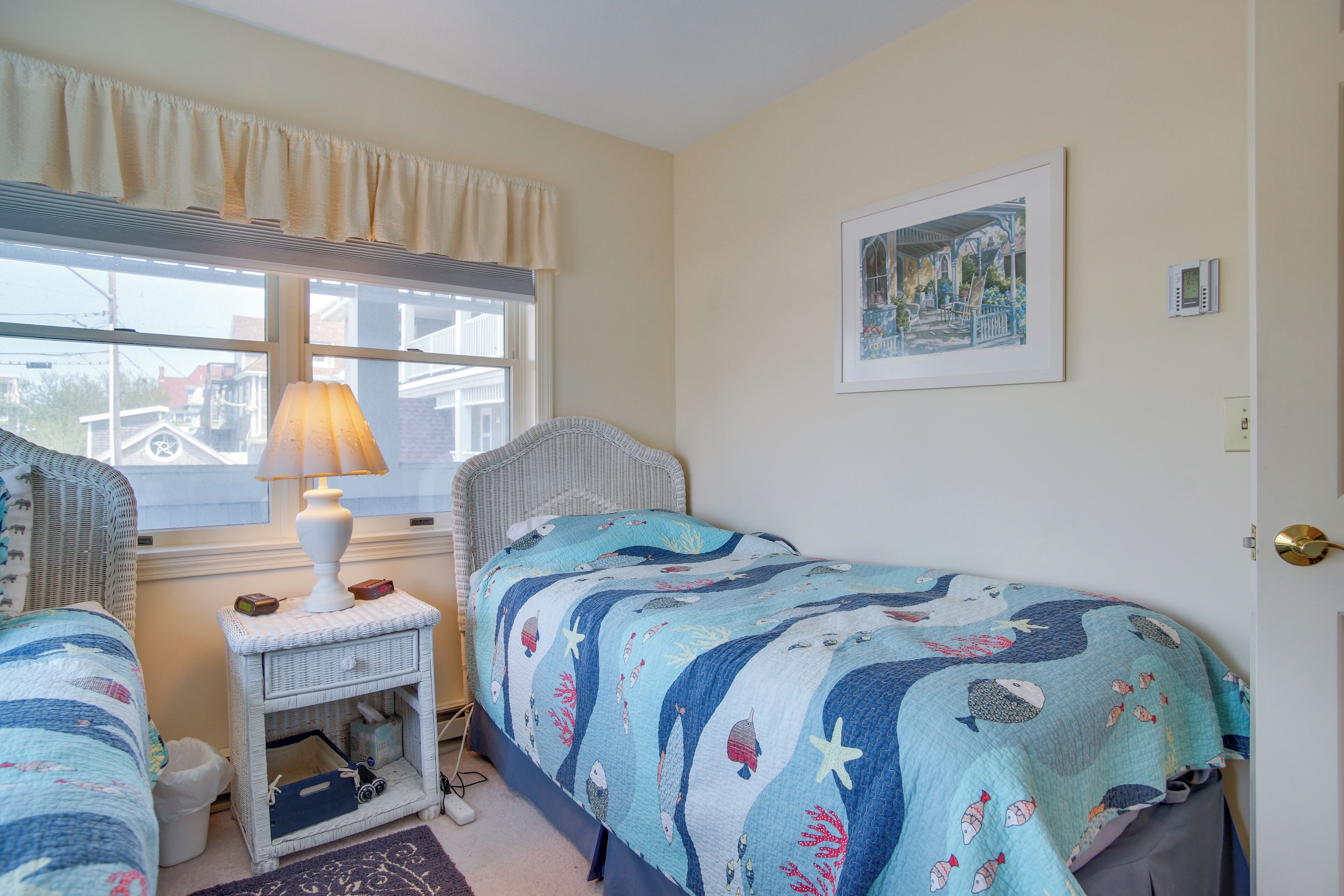 A bedroom with two beds, a bedside table with a lamp, a framed picture on the wall, and a window with curtains.
