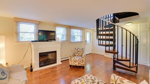 A spacious living room with hardwood flooring, a fireplace, a television, a patterned armchair, a staircase, and a door leading to another room. - Shop, Dine, Explore Commercial St! Coastal Retreat (Provincetown)