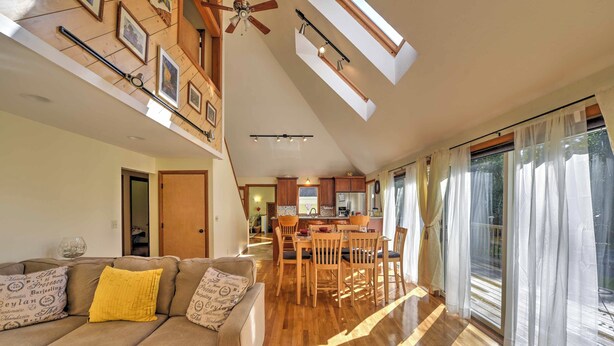 A spacious living area with a high ceiling, wooden flooring, a sofa with patterned cushions, a dining table with chairs, and a kitchen in the background.